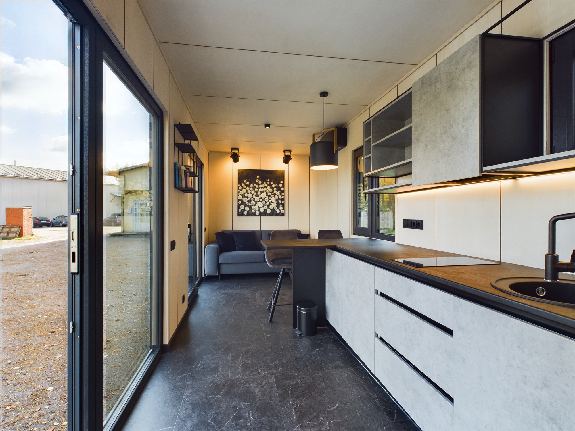 Moderna, gaiša virtuve ar tumšu virsmu un atvērtu skatu uz viesistabu. Kvalitatīva nekustamā īpašuma fotogrāfija.