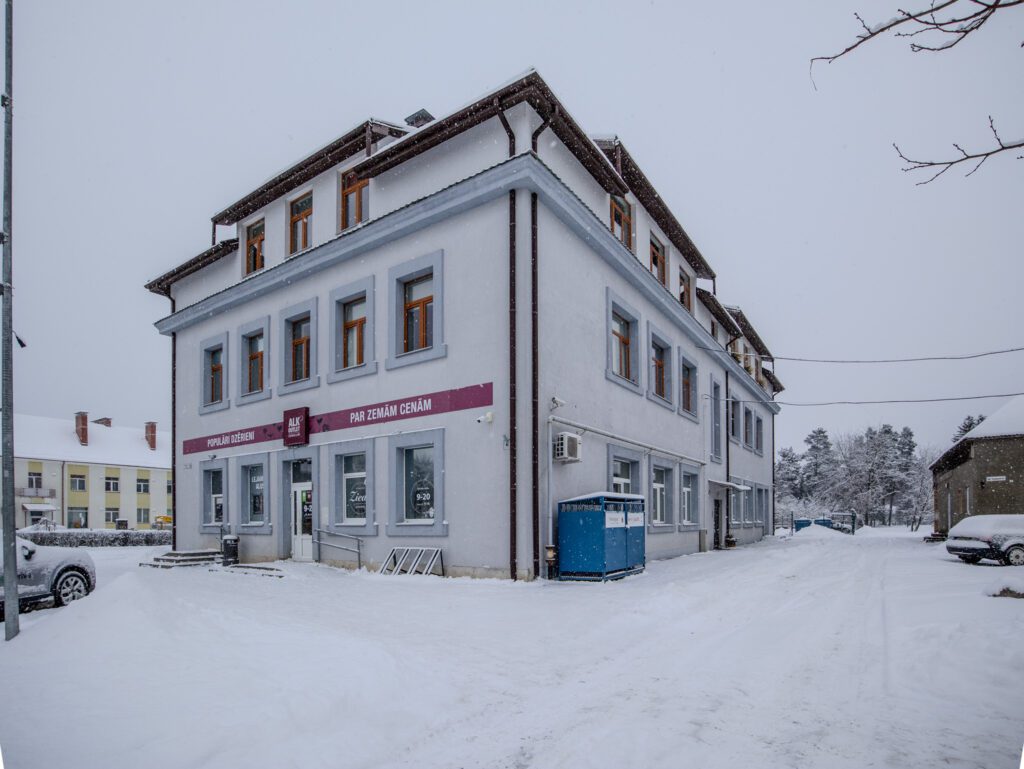 Baltas, divstāvu komercīpašuma ēkas fasāde ziemas ainavā (Vangaži). Kvalitatīva komerctelpu fotogrāfija.