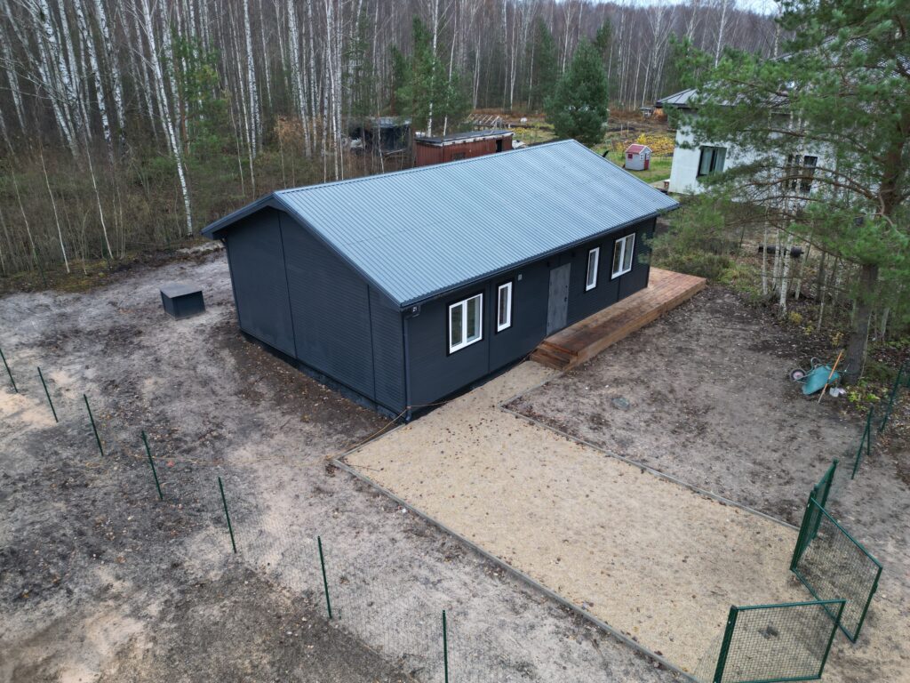 Drona kadrs: Melna vai tumši pelēka moduļu māja ar baltiem logiem, kas uzstādīta uz zemes gabala. Kvalitatīva nekustamā īpašuma pārdošanas fotogrāfija.