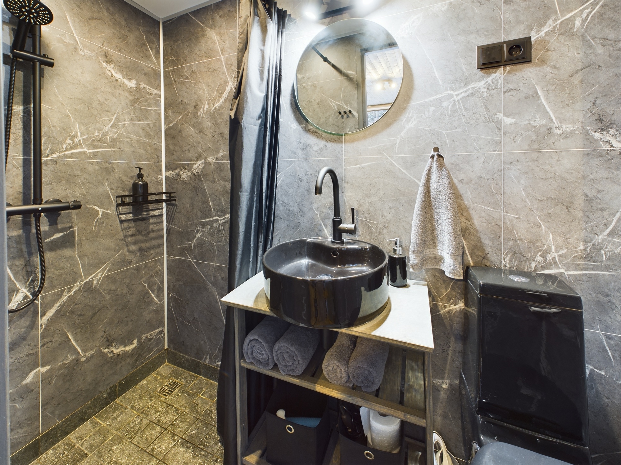 Tuvplāns: Stilīgs sanmezgls ar melnu izlietni, tumšu dušas stieni un apaļu spoguli. Kvalitatīva vannas istabas fotogrāfija.