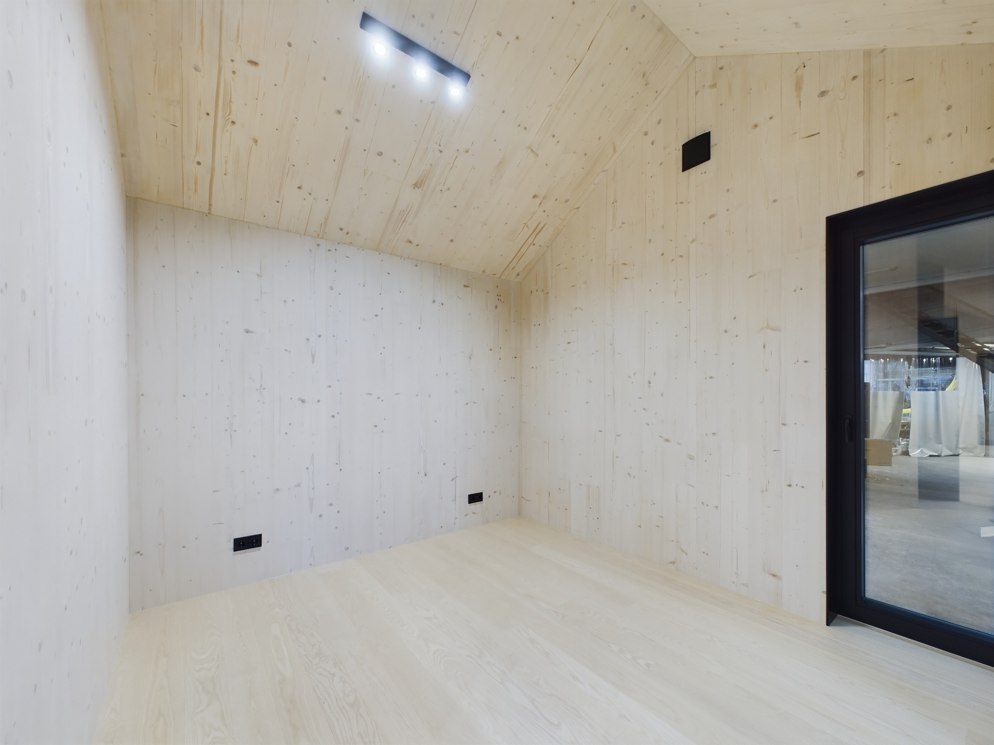 Tīra, tukša moduļu mājas telpa ar gaišu koka apdari un lielu melni kadrētu logu. Kvalitatīva arhitektūras fotogrāfija.