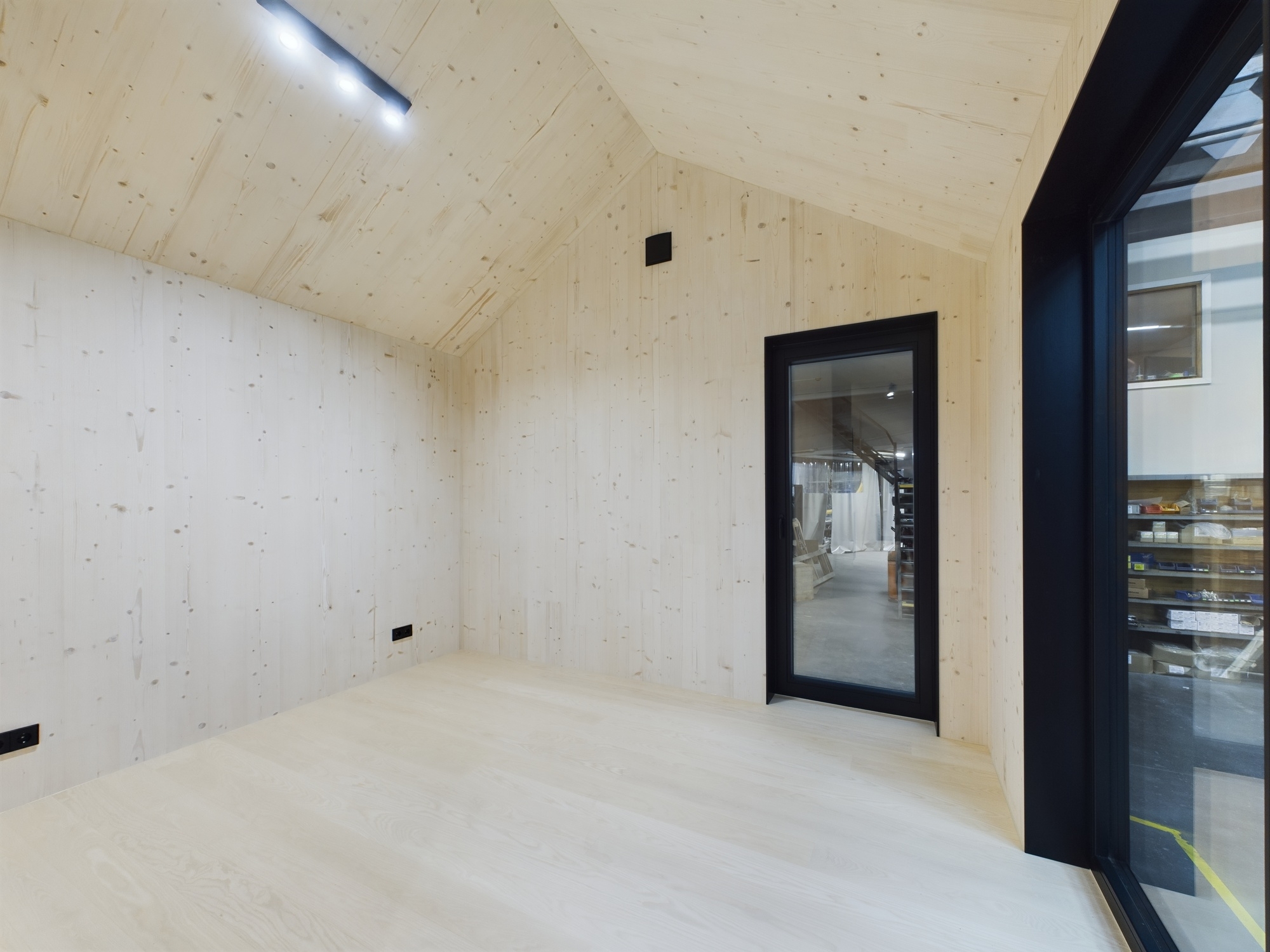 Tīra, tukša moduļu mājas telpa. Interjers ar gaišu koka apdari un lielu melni kadrētu logu. Kvalitatīva arhitektūras fotogrāfija.