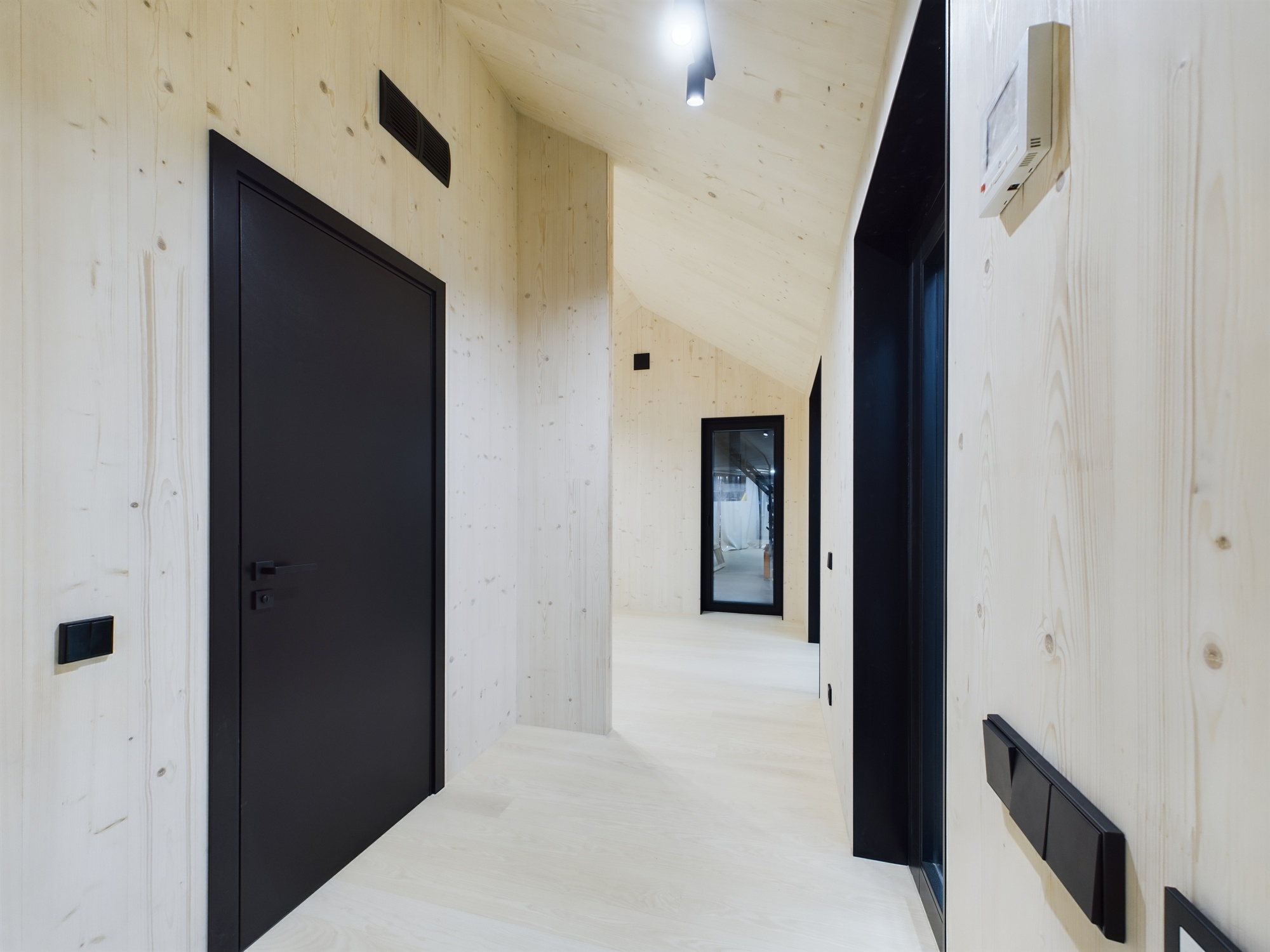 Tīras līnijas. Koridors ar gaišiem koka paneļiem un melnām, mūsdienīgām iekšdurvīm. Kvalitatīva moduļu mājas fotogrāfija.
