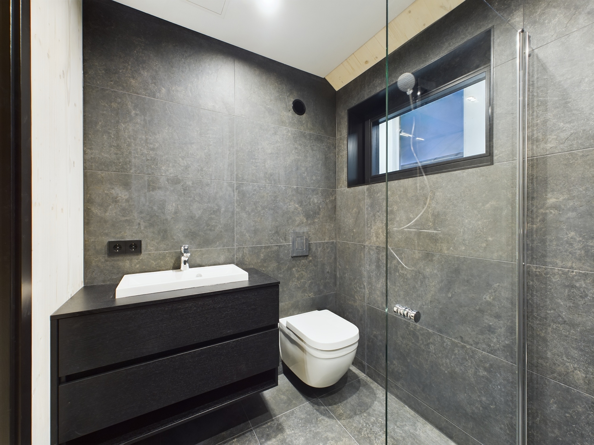 Augstas klases sanmezgls ar tumšām flīzēm, iebūvētu dušu un modernu izlietni. Kvalitatīva interjera fotogrāfija.