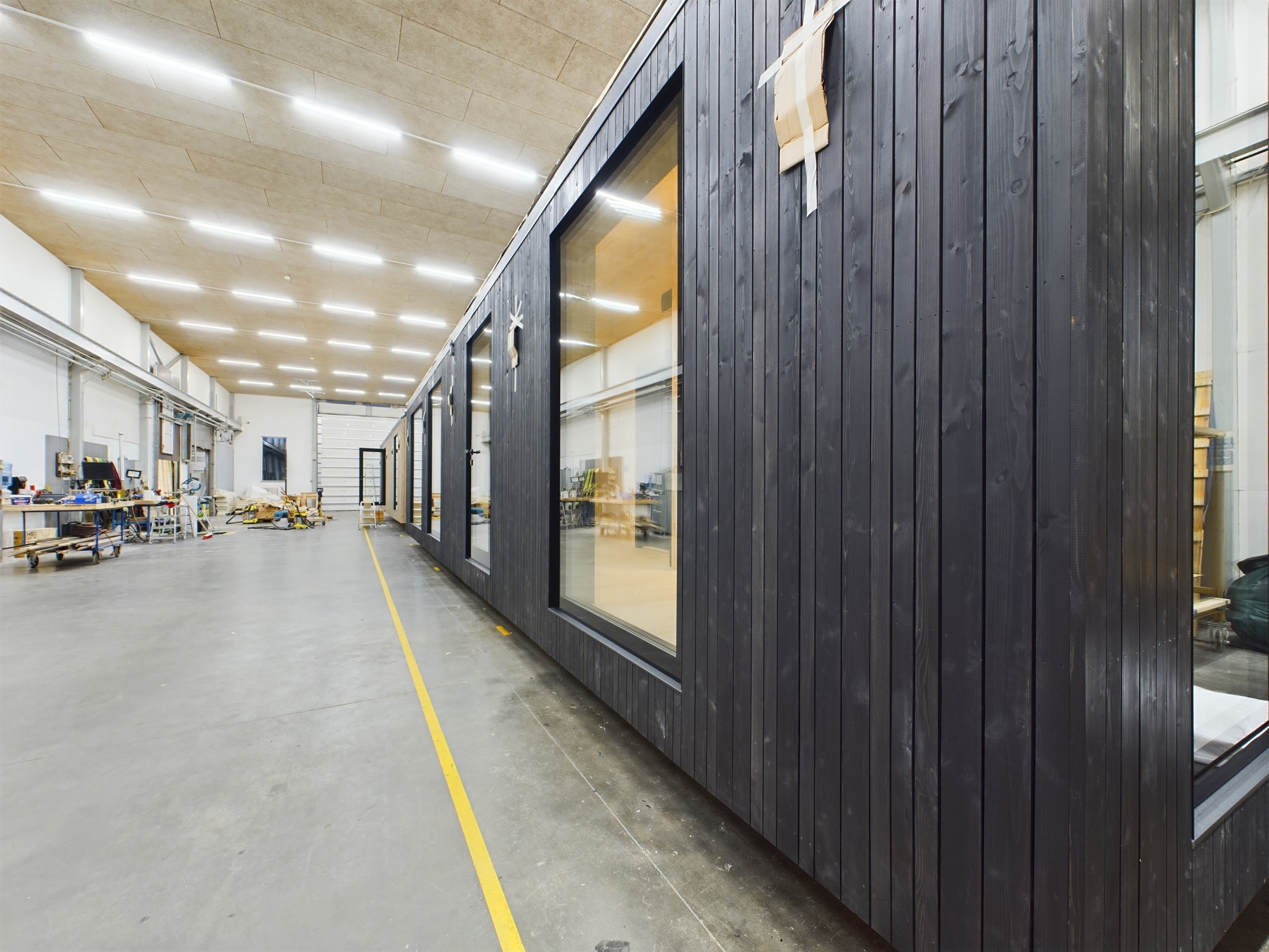 Moduļu mājas fasāde ar melnu apdari un logiem, kas uzņemta ražošanas telpās. Kvalitatīva būvniecības fotogrāfija.