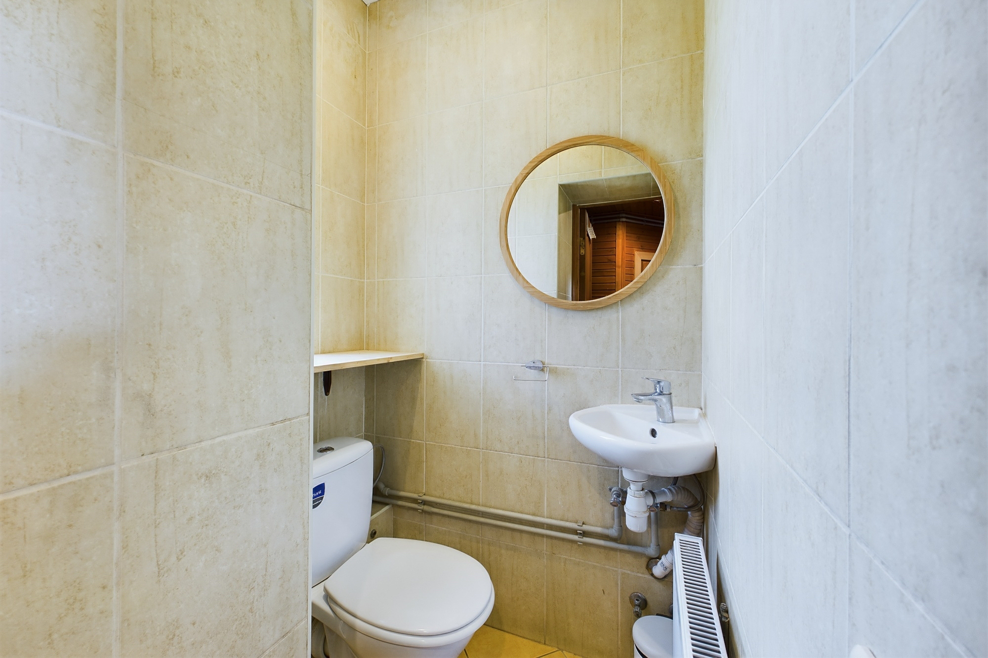 Tīra, kompakta tualetes telpa ar nelielu izlietni un apaļu spoguli. Kvalitatīva interjera fotogrāfija.